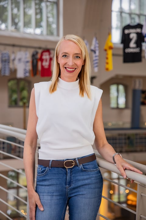 Blonde woman in white sleeveless shirt and blue jeans smiling at camera in indoor mall setting